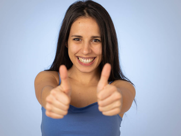 Portrait of beautiful young latin woman showing thumbs up sign feeling cheerful, happy and satisfied. Young happy student woman making thumb up in joyful approval gesture isolated on blue background. - Zdjęcie, obraz