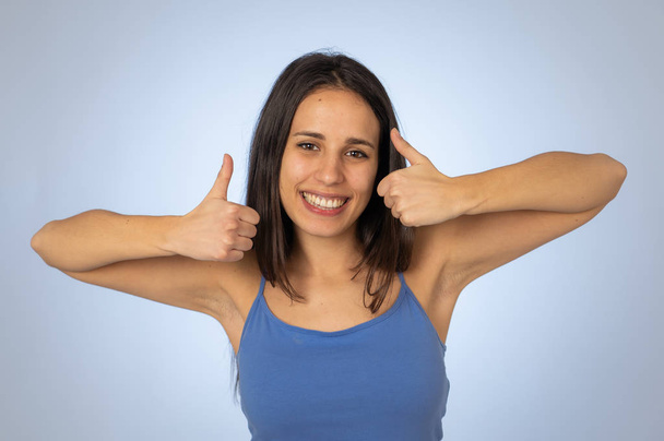 Portrait of beautiful young latin woman showing thumbs up sign feeling cheerful, happy and satisfied. Young happy student woman making thumb up in joyful approval gesture isolated on blue background. - Фото, изображение
