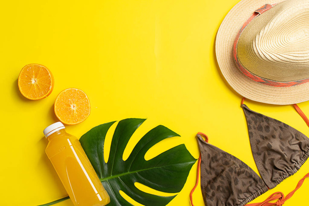 Zomer strand accessoires Hat badpak fles oranje sap Bright gele achtergrond grote groene bladeren tropische plant. - Foto, afbeelding