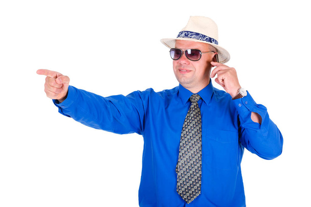 Успешный и улыбающийся бизнесмен в синей рубашке, галстуке, солнцезащитных очках и белой шляпе. Говорит по мобильному телефону и показывает указательный палец. Изолированный белый фон, концепция лидерства
 - Фото, изображение