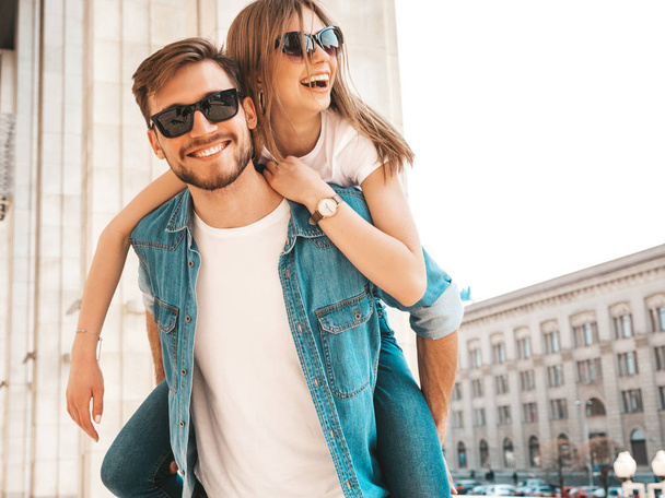 Smiling beautiful girl and her handsome boyfriend in casual summer clothes. Man carrying his girlfriend on the back and she raising her hands.Happy cheerful family having fun on the street background  - Fotoğraf, Görsel