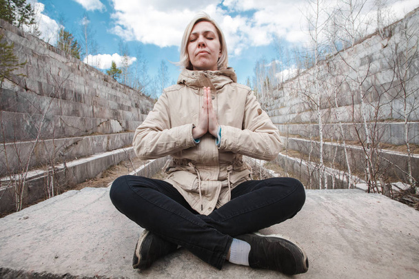Portrait of a meditating blonde outdoors on the background of a marble quarry. Unity with the concept of nature. Girl sitting on a stone in a windbreaker. - Фото, изображение