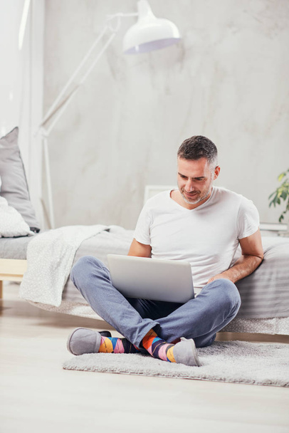 Przystojny mężczyzna w średnim wieku siedzi na podłodze, opierając się na łóżku i za pomocą laptopa. - Zdjęcie, obraz
