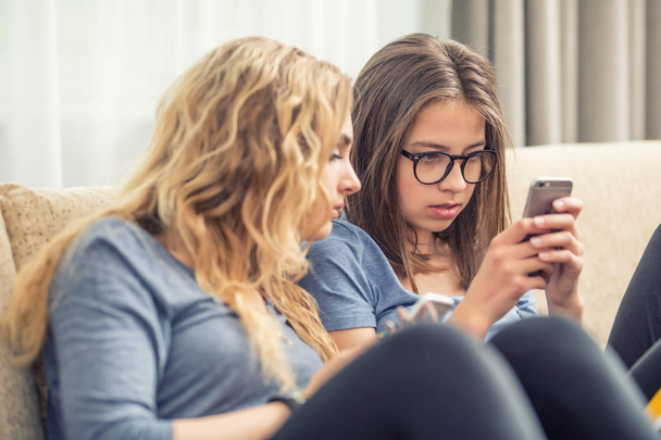 Kaksi teini girs tai sisaret käyttävät älypuhelimia olohuoneessa
 - Valokuva, kuva