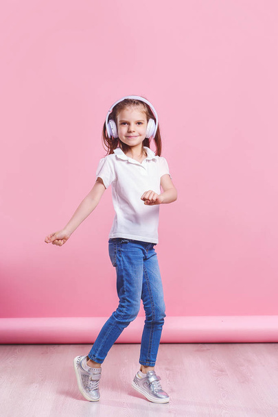 Girl listening to music in headphones an dance on pink background. Cute child enjoying happy dance music, close eyes and smile posing on studio background wall - Photo, Image