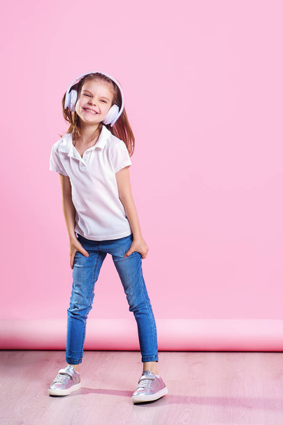 Girl listening to music in headphones an dance on pink background. Cute child enjoying happy dance music, close eyes and smile posing on studio background wall - Foto, immagini