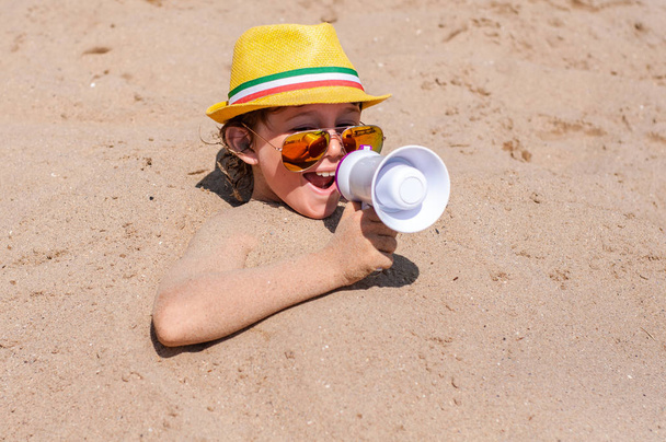 Cheerful boy in sunglasses and a hat with a mouthpiece in the hands immersed in the sand on the beach. Announces beach season open - Photo, Image