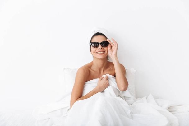 Beautiful amazing woman with towel on head lies in bed under blanket isolated over white wall background wearing sunglasses. - Valokuva, kuva