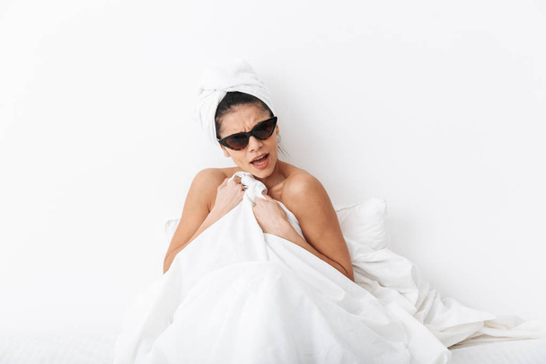 Displeased woman with towel on head lies in bed covering body under blanket isolated over white wall background wearing sunglasses. - Valokuva, kuva