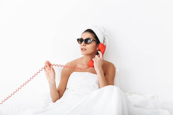 Emotional woman with towel on head lies in bed under blanket isolated over white wall background wearing sunglasses talking by phone. - Zdjęcie, obraz