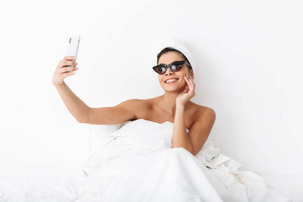 Beautiful emotional woman with towel on head lies in bed under blanket isolated over white wall background wearing sunglasses take a selfie by mobile phone. - Foto, Bild