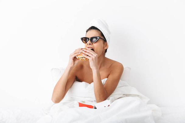 Emotional woman with towel on head lies in bed under blanket isolated over white wall background wearing sunglasses eat burger. - Valokuva, kuva