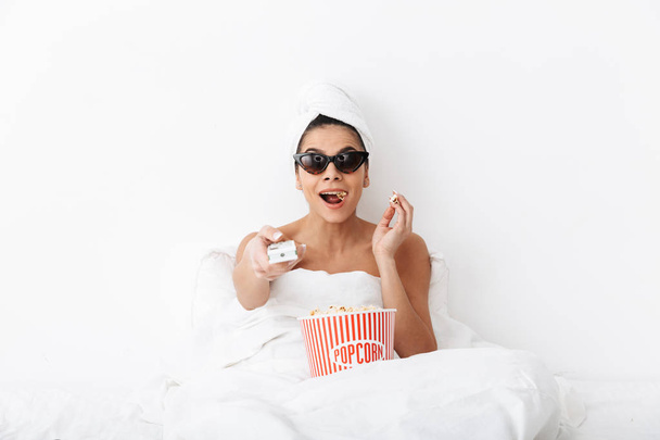 Happy excited woman with towel on head lies in bed under blanket isolated over white wall background wearing sunglasses eat popcorn watch film tv. - Photo, image