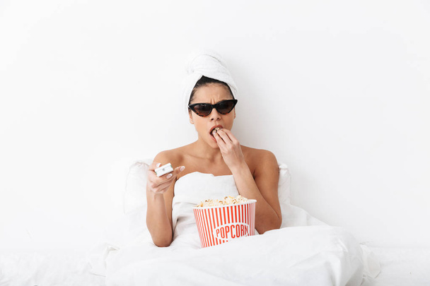 Beautiful concentrated woman with towel on head lies in bed under blanket isolated over white wall background wearing sunglasses eat popcorn watch film tv. - Foto, immagini