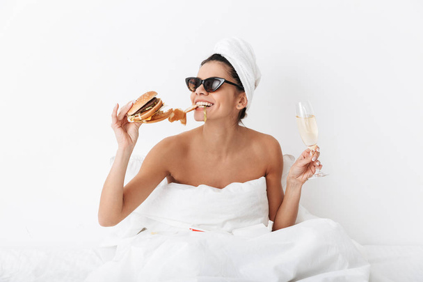 Emotional woman with towel on head lies in bed under blanket isolated over white wall background wearing sunglasses eat burger. - Foto, imagen