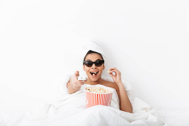 Başında havlu ile Mutlu heyecanlı kadın beyaz duvar arka plan üzerinde izole battaniye altında yatakta yatıyor güneş gözlüğü giyen patlamış mısır izlemek film tv yemek. - Fotoğraf, Görsel