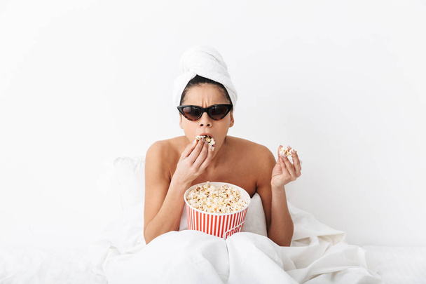 Scioccato donna spaventata con asciugamano sulla testa si trova a letto sotto coperta isolato su sfondo muro bianco indossare occhiali da sole mangiare popcorn guardare la tv film
. - Foto, immagini
