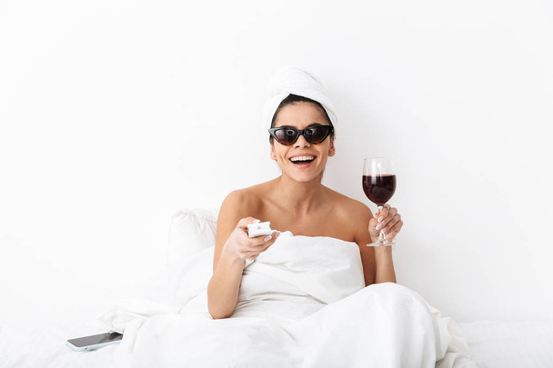 Woman with towel on head lies in bed under blanket isolated over white wall background wearing sunglasses drinking wine watch tv film. - Foto, Imagem