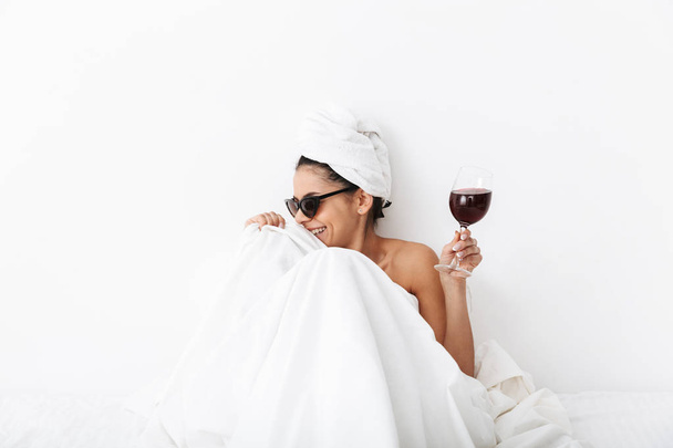 Laughing happy woman with towel on head lies in bed under blanket isolated over white wall background wearing sunglasses drinking wine. - Foto, afbeelding