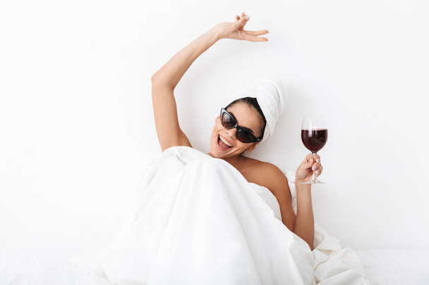 Beautiful laughing happy woman with towel on head lies in bed under blanket isolated over white wall background wearing sunglasses drinking wine. - Foto, imagen