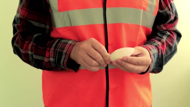 Man electrician dressed in a reflective vest give to the camera energy saving LED lamp. Hands closeup isolated on light green background. - Footage, Video