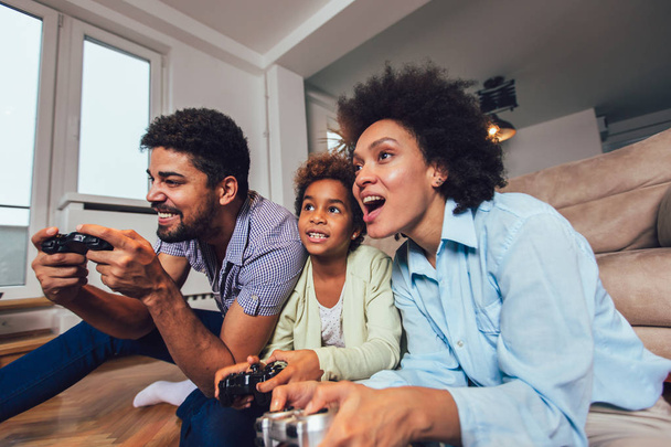 Hymyilevä perhe istuu sohvalla yhdessä pelaamassa videopelejä
 - Valokuva, kuva