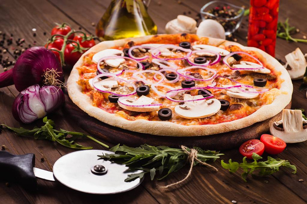 Italian pizza with mushrooms, olives and onion rings with ingredients on wooden table (close) - Photo, Image
