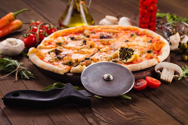 delicious Italian seafood pizza with ingredients on a dark wooden table (close). Cut one piece - Photo, Image