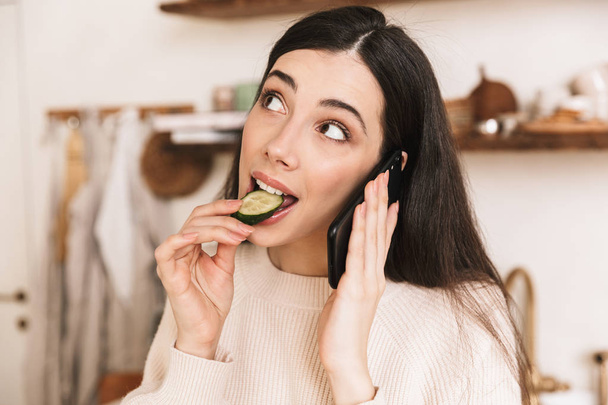 Picture of adorable brunette woman 30s eating cucumber while tal - Fotografie, Obrázek