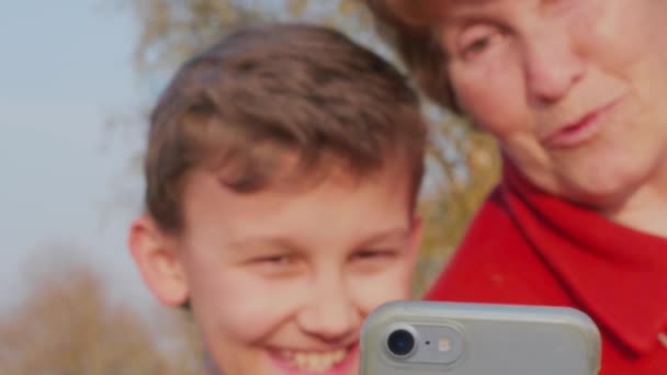 笑顔の先輩女性とティーンエイジャーの男の子は、外のモバイルで自分撮りを取って、電話、感情に焦点を当てます。長老の女性と子供は、屋外の携帯電話で自分撮りを取得します。スマートフォンを使って写真を撮る祖母や孫、一日。高齢者と子供のアクティブなライフスタイル - 映像、動画