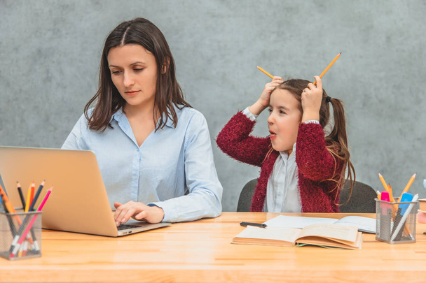 Daughter and mother on a gray background. During this, the girl puts two yellow crayons on her head, showing the tongue. Mom looks seriously while working on a laptop. - Fotografie, Obrázek