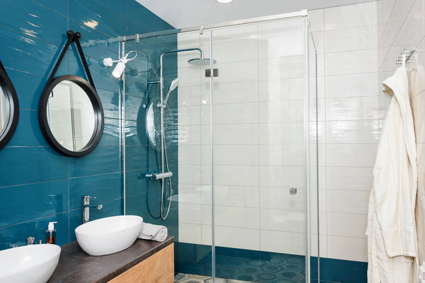 Moderno cuarto de baño interior con una estantería de madera, dos lavabos de pie en él y espejos redondos. Cabina de ducha de cristal transparente, baño escandinavo minimalista en apartamento loft
 - Foto, Imagen