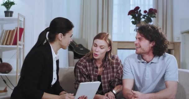 Charismatic couple have a discussion with a real estate agent representing the house plan at the end she make a great deal with her client shaking hands very impressed while sitting on the sofa - Materiał filmowy, wideo