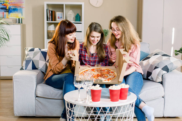 Tre amiche che cenano a casa. Foto di allegre ragazze sorprese che celebrano una festa a casa, aprendo scatola di cartone con con pizza
. - Foto, immagini