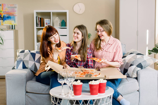 Portrait of three happy young girlfriends eating pizza on sofa at home. Beautiful ladies smiling and communicating about every day life: work, home, men. - Foto, imagen