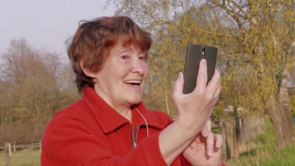 外で携帯でセルフィーを撮る笑顔の先輩女性。おばあさんは、屋外で携帯電話で自分撮りを取得します。スマートフォンを使って写真を撮るおばあちゃん、一日。高齢者のアクティブなライフスタイル。現代の技術を使用しておばあちゃん - 映像、動画