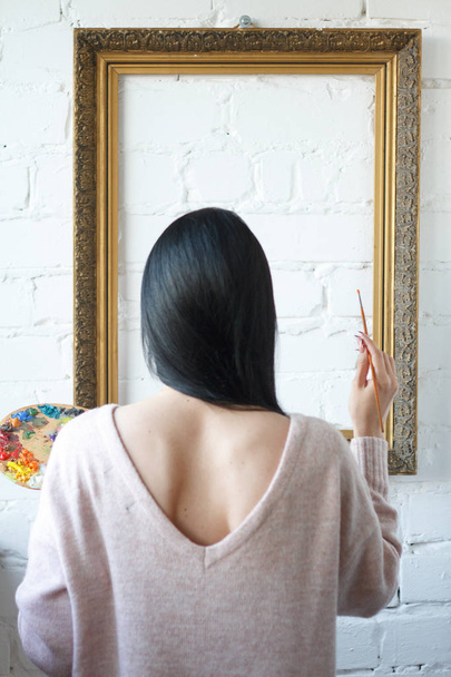 Uzun siyah saçlı genç bir kadın boş bir vintage çerçeve nin arka plan üzerinde renkli boyalar ile bir fırça ve palet tutarak sırtını tutuyor - Fotoğraf, Görsel