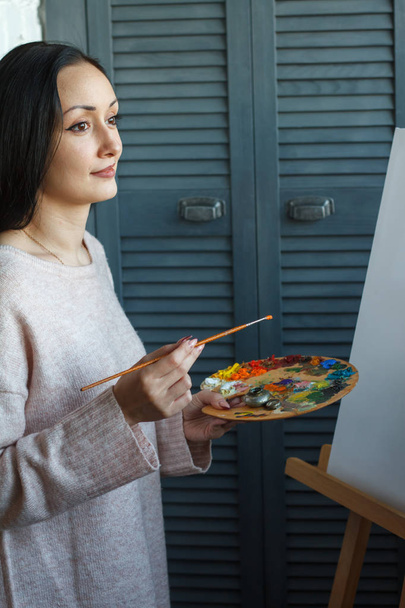 A young woman of eastern appearance with long black hair holds a brush and a palette with multi-colored acrylic paints in her hands - Photo, Image