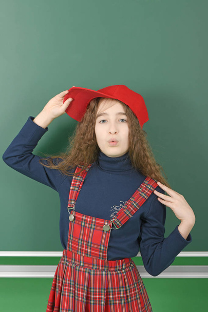 Школярка біля зеленої шкільної дошки. Молода грайлива дівчина тримає червону шапку
.  - Фото, зображення