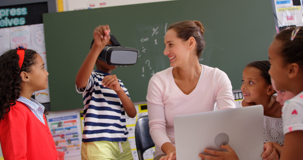 Front view of African american schoolboy using virtual reality headset with teacher and classmates in the classroom. Female teacher using laptop  - Footage, Video