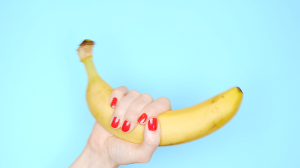kobiece ręce z czerwonymi gwoździami trzymają żółty banan na niebieskim tle - Materiał filmowy, wideo