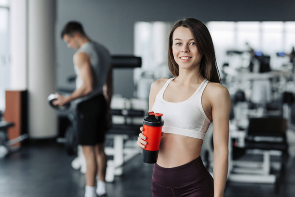 Atractiva chica deportiva sonriendo y bebiendo agua mientras está de pie en el gimnasio con el niño entrenando en segundo plano
. - Foto, imagen