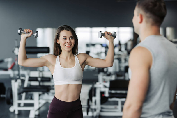 Young couple is working out at gym. Attractive woman and handsome muscular man trainer are training in light modern gym. Beautiful girl squats with kettlebell under the supervision of the coach. - Photo, Image