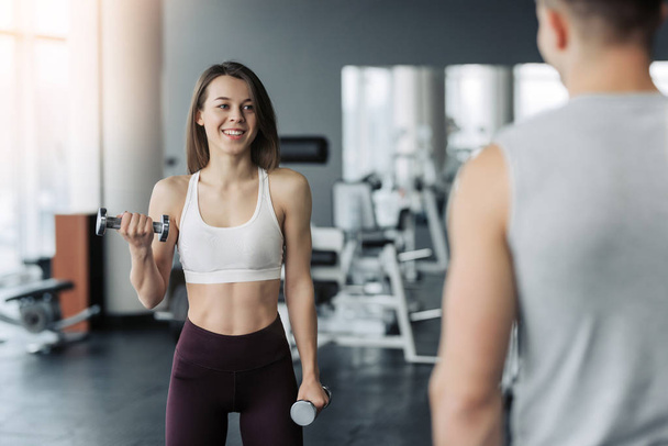 Young beautiful woman doing exercises with dumbbell in gym. Glad smiling girl is enjoying with her training process. She is working hard. Her trainer looking at this process. - Photo, Image