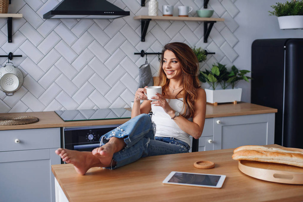 Chica alegre en camiseta blanca usando tableta mientras se relaja en una silla en casa en la acogedora cocina
. - Foto, imagen