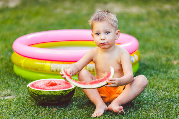 Téma letní sezónní potraviny šťavnatý meloun bobule. malé funny boy dítě dva roky sedí dvorku zelené trávě, trávník a jíst meloun kus drží za ruce. Nahý v šortkách a naboso - Fotografie, Obrázek