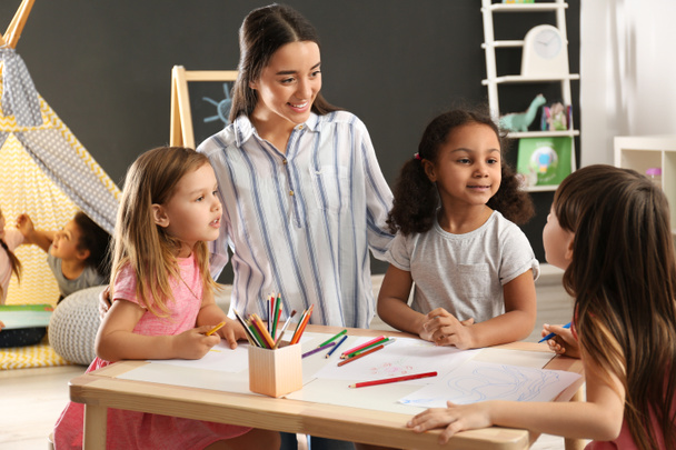 Cute little children with nursery teacher drawing at table in kindergarten. Indoor activity - Zdjęcie, obraz