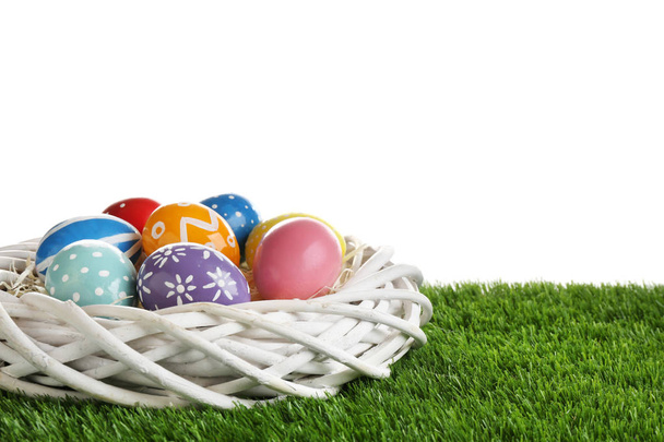 Wicker nest with painted Easter eggs on green grass against white background, space for text - Фото, зображення