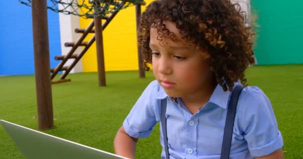 Front view of mixed-race boy studying on laptop in school playground. She is sitting on grass 4k - Materiał filmowy, wideo