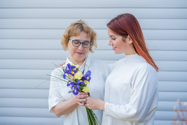 Adult daughter gives flowers to her mother outside, in the courtyard of the house. Spending time together, celebrating at home on weekends. Mothers Day. Warm intergenerational relationships - Foto, Imagen